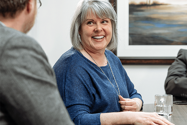 A smiling older woman with gray hair and a blue sweater sits at a table, talking to a man in glasses. A glass of water and a framed landscape painting are visible in the background.