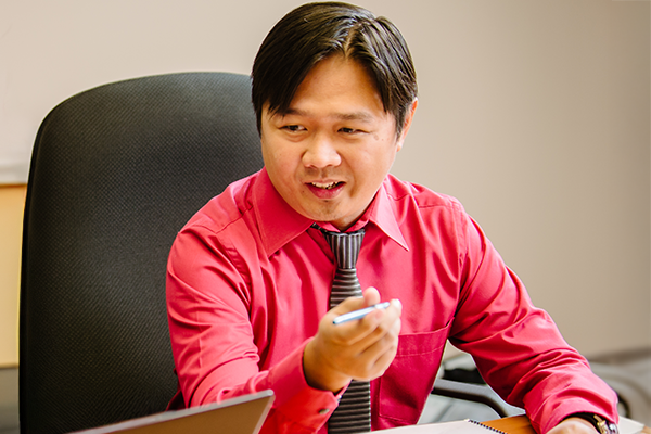 A man in a red dress shirt and striped tie sits at a desk, holding a pen and discussing the functionality similarities and differences between Dynamics GP and Dynamics 365, appearing engaged in the meeting.